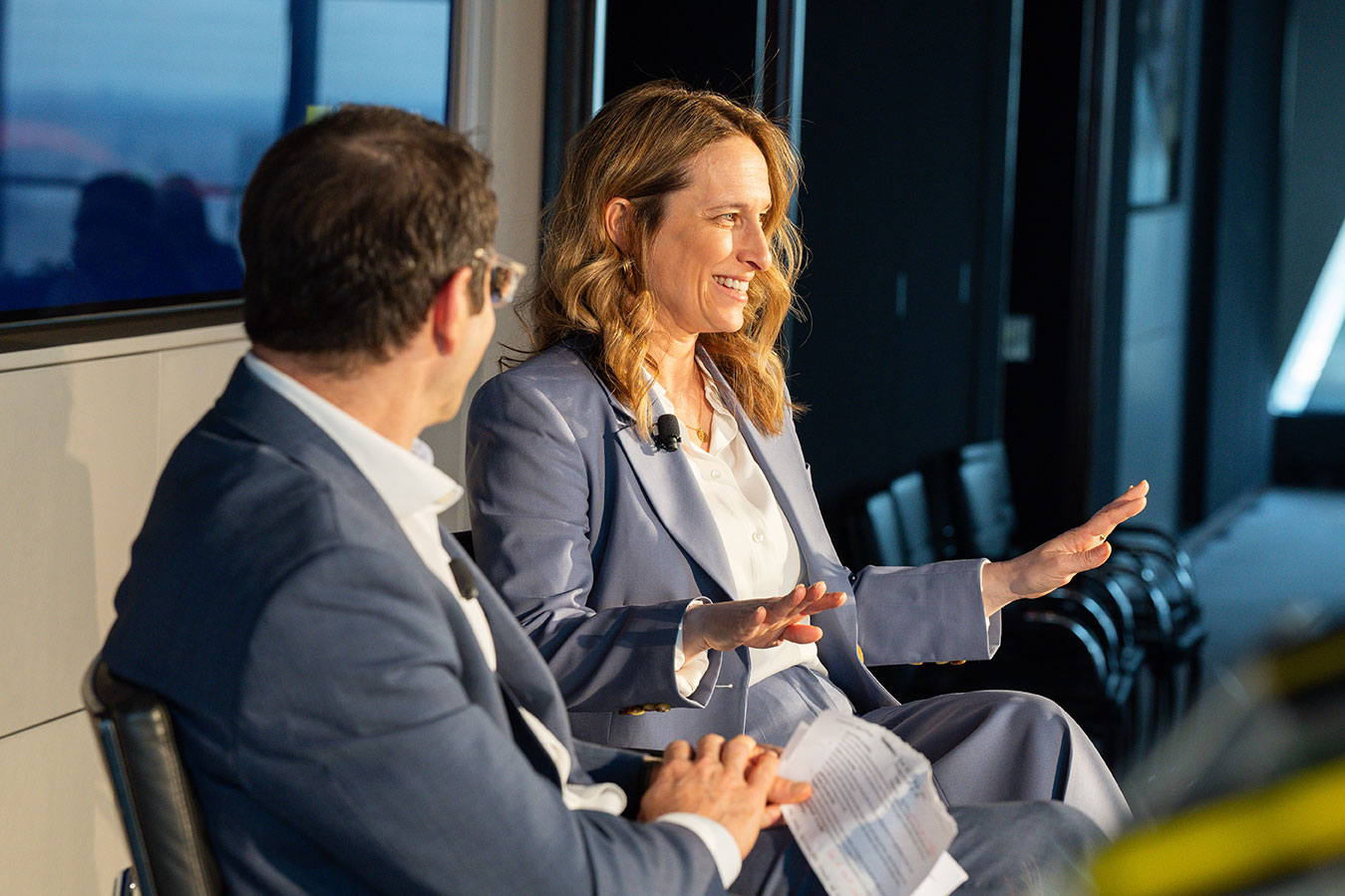 Two panelists in conversation at Leadership Now 2025 summit, city skyline visible through windows behind them